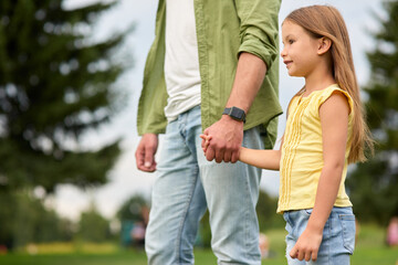 Obraz premium Side view of cute little girl and her father holding each other hands, standing in a green park on a warm day, walking together outdoors