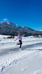 Fototapeta na wymiar A woman in colorful outfit skating on a frozen Weissensee lake in Austria. The lake is surrounded by mountains. The ice rink is well prepared. Winter activity. Winter wonderland. Happiness
