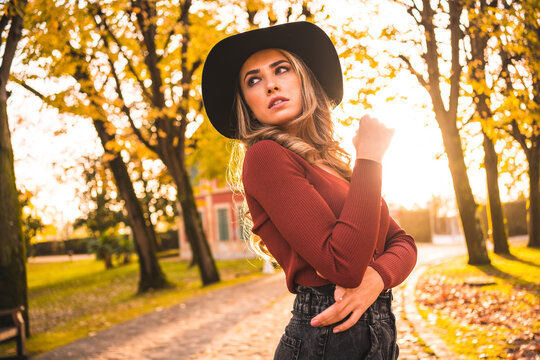 Autumn lifestyle at sunset, blonde Caucasian girl in a red sweater and black hat, enjoying nature in a park with trees