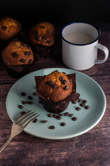 Muffins with pieces of chocolate, cup of milk and plate with chocolate chips on the wooden table.