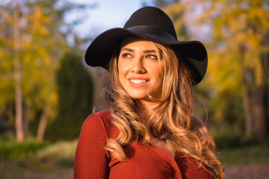 Autumn lifestyle, blonde Caucasian girl in a red sweater and a beautiful black American hat, enjoying nature in a park with large trees