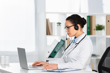Fototapeta na wymiar Side view of confident African American doctor in headset looking at laptop, while sitting at desk on blurred background