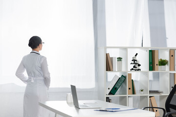Back view of african american doctor standing near window with blurred workplace on foreground