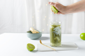 Woman hand squeezing lime into glass with ice green matcha latte on white background.