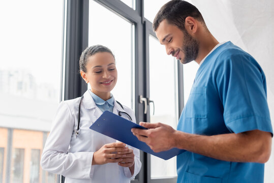 Smiling nurse holding clipboard near african american doctor in clinic