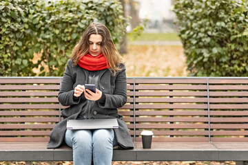 Young woman holds a smartphone and checks messages while sitting on a park bench with a laptop on...