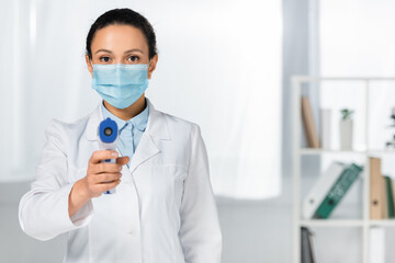 African american doctor in medical mask holding infrared thermometer on blurred foreground in clinic