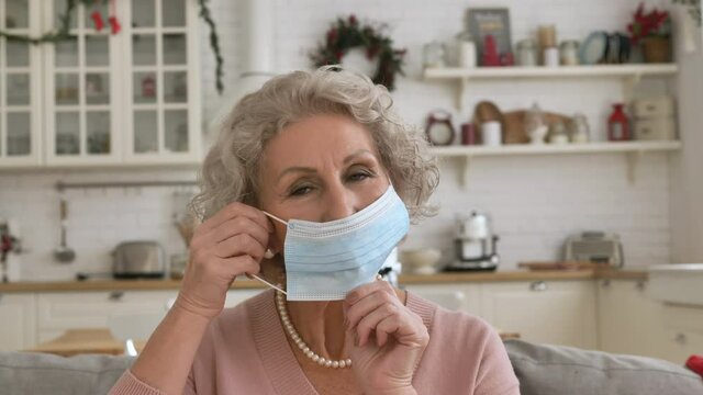 Concentrated grandmother with trendy grey hairstyle puts on protective facial mask against kitchen furniture closeup slow motion