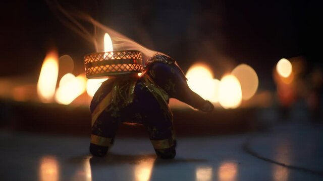 small elephant incense holder with smoke coming out and an out fo focus background with diya oil lamps lit behind it with flickering flames showing the hindu festival of diwali