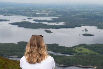 Panoramic view of the Irish countryside with trees, green vegetation and lakes with mountains and hills, cloudy day in Ireland. Girl sitting on the edge