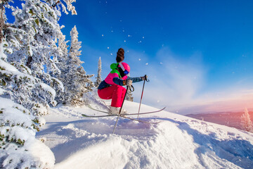 Woman skier jumping in pink skis on background of blue sky and snowy forest in mountains, dust from snow