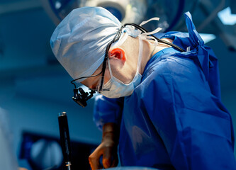 Neurosurgeon in blue scrubs and surgical glasses provides operation in up to date operation room...