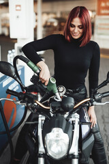 Naklejka na ściany i meble A modern gas station and attractive female driver with red hairs. A girl fueling own bike with petrol. Vintage light photo.