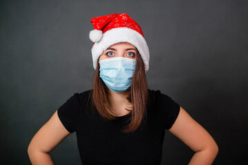 Portrait of a woman in a black T-shirt, a disposable medical mask and a Christmas hat on a gray background. Alone quarantine christmas celebration concept.