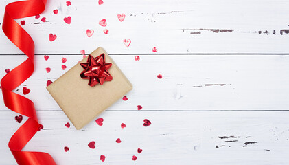 Gift on a white wooden background with a red bow, scattered red hearts, satin ribbon. Valentine's day. Top view, free space for text, flatly.
