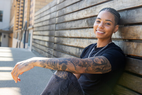 Young African American woman with tattoos on arm wearing black tee shirt sitting against wood wall outside
