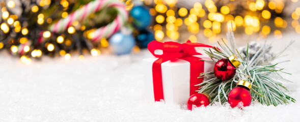 Present with red ribbon, pine or spruce branch with red balls on the snow on the background of blurred Christmas tree and lights of garland.