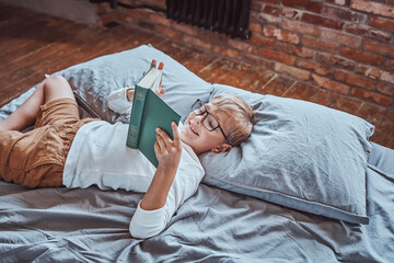 Dressed in casual clothing cheerful and smiley boy reads book lying down on a bed in living room.