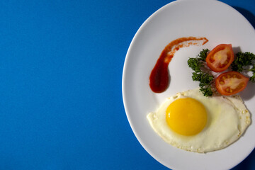 A dish of fried eggs. Blue background with space for text. Bachelor's Breakfast. Fried eggs on a white plate with tomatoes, herbs, ketchup