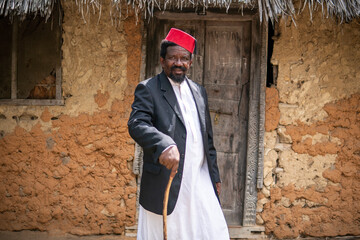 An African Older Man in Red Muslim Taqiyyah Fez Hat And Blazer Walking with a stick for limping people Near the Basic Hut with Thatched roof in Small Remote Village in Tanzania, Pemba island, Zanzibar - obrazy, fototapety, plakaty