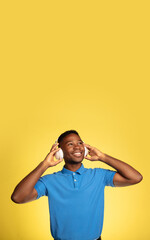 Music in headphones. Young african-american man's portrait isolated on yellow studio background, facial expression. Beautiful male model, copyspace. Concept of human emotions, sales, ad. Flyer