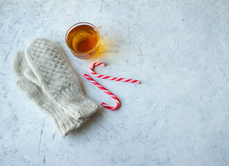 winter fluffy warm knitted mittens, a glass mug with tea and Christmas candies lie on a gray-white textured background, top view, festive minimalism