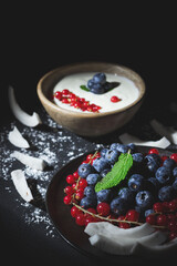 Black plate with red berries and blueberries, peppermint leaf on black background with bowl of blueberries and yogurt in the background