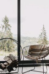 Modern chair with cozy blanket and firewood on metal stand at window view on mountain in cabin