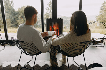Stylish couple in sweaters with warm tea relaxing at modern fireplace with view on mountains