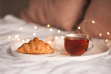 Mug of fresh black tea staying on white tray with tatsy cake over glowing Christmas lights close up. Good morning.