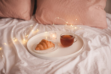 Cup of black tea with fresh tasty croissant staying on white tray over Christmas lights in bed close up. Good morning. Breakfast time.
