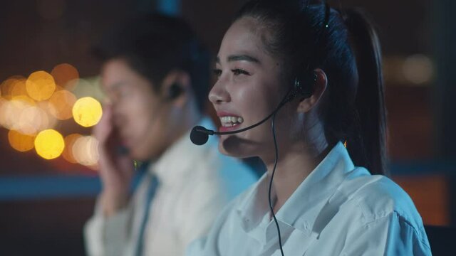 Millennial Asia young call center team or customer support service executive using computer and microphone headset working technical support in late night office. Telemarketing or sales job concept.