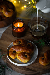 Pouring milk into glass with coffee, standing on wooden tray with two Swedish saffron buns (lussekatter) and lit  candle .