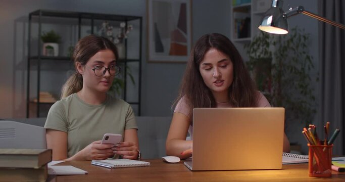 Portrait of Caucasian twin sisters sitting at desk in cozy room with laptop and smartphone. Female tapping on computer and talking with twin sister who is texting on cellphone. Indoors concept