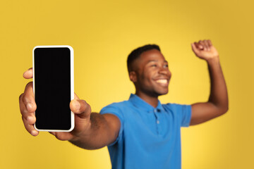 Shows phone screen, winner. Young african-american man's portrait isolated on yellow studio background, facial expression. Beautiful male model, copyspace. Concept of human emotions, sales, ad.