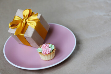 Gift box wrapped by kraft brown paper with beautiful yellow satin ribbon and small sweet cake on pink plate background. Universal gifts design for all kind of celebration. Sweet bakery food.