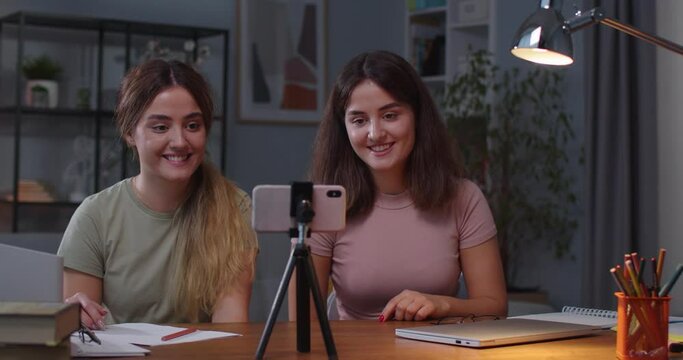 Caucasian happy beautiful young female twins waving hands and smiling while talking on online call on smartphone. Portrait of joyful sisters having video chat on cellphone at cozy home Leisure concept