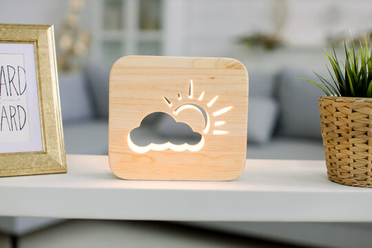 Close up cropped image of decorative wooden night lamp with sun and cloud picture, on light table with flower pot and photo frame. Cozy living room interior with gray sofa on the background