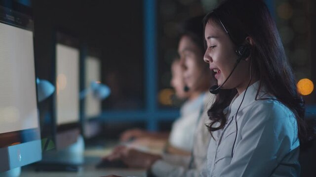 Millennial Asia young call center team or customer support service executive using computer and microphone headset working technical support in late night office. Telemarketing or sales job concept.