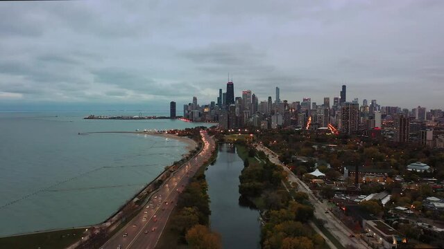 Beautiful panning out aerial view of the Chicago skyline over the South Lagoon in Lincoln park by Lake Michigan with Traffic and city lights beginning to turn on just before sunset on a cloudy evening