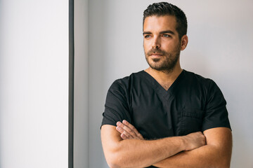 Confident male dentist in black medical scrubs standing with arms crossed against wall