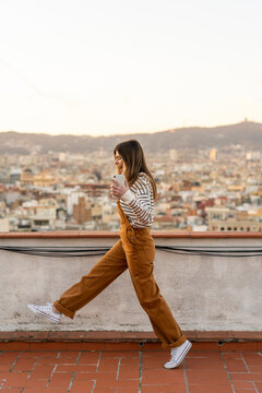 Happy young woman with smartphone walking on roof terrace