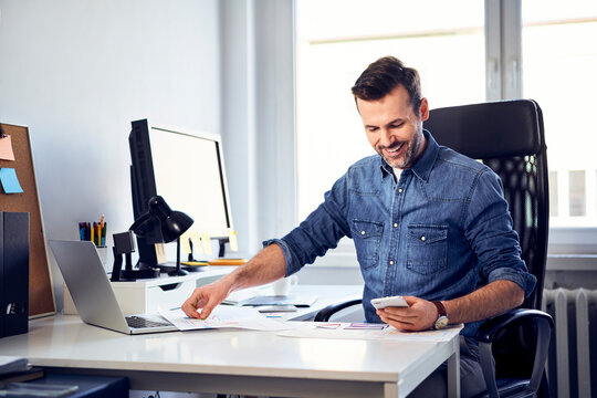 Smiling man with smartphone and draft working at desk in office