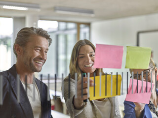 Happy colleagues discussing with sticky notes at glass pane