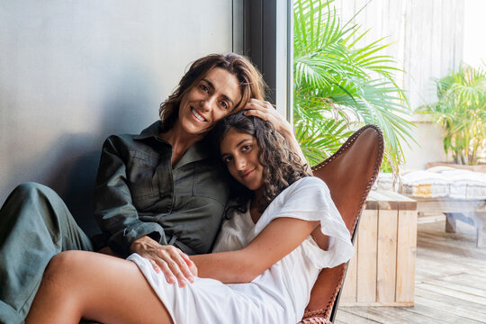 Smiling mother and daughter embracing while sitting on chair at home