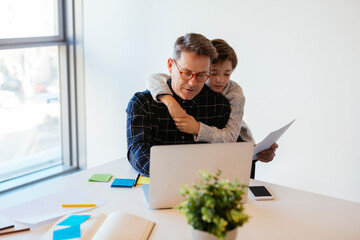 Businessman using laptop at desk in office with son embracing him