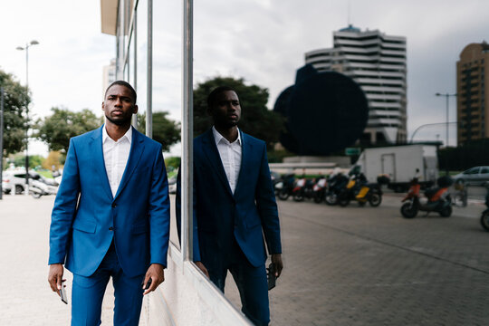 Confident male African entrepreneur walking by reflection of himself on window in city