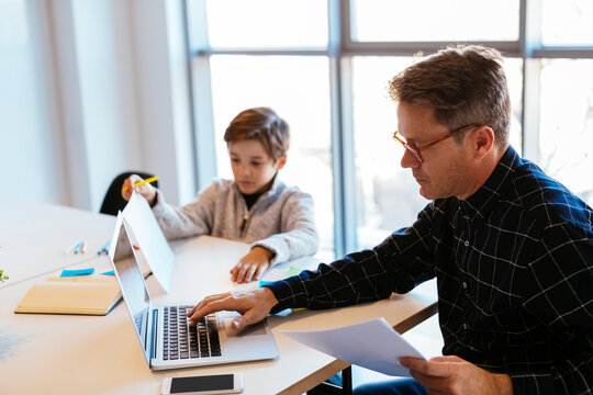 Businessman using laptop at desk in office with son sitting next to him