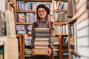 Portrait of a beautiful smart 20s female student in glasses holding stack of books in library or bookshop