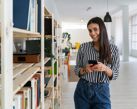 Smiling Beautiful Young Female Architect Holding Smart Phone While Standing By Rack At Creative Office